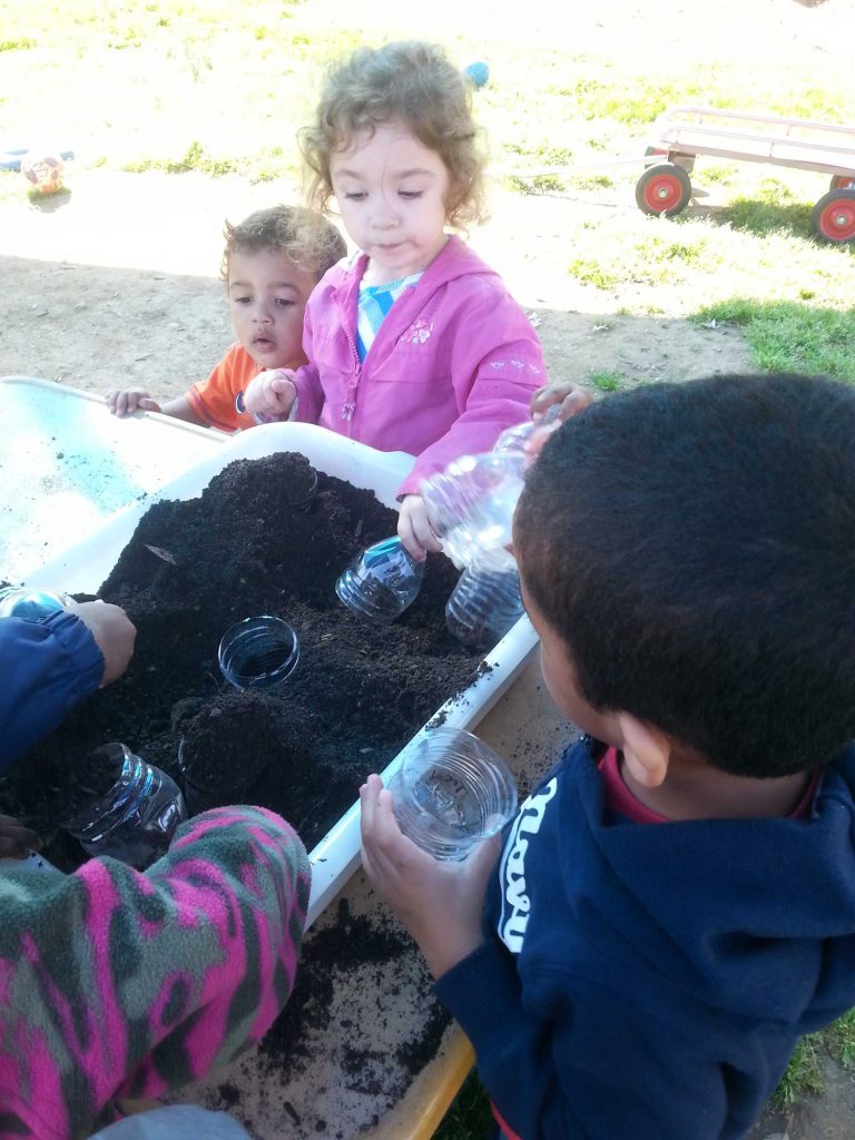 A group of children engaged in outdoor planting activities, with black soil and half-cut water bottles