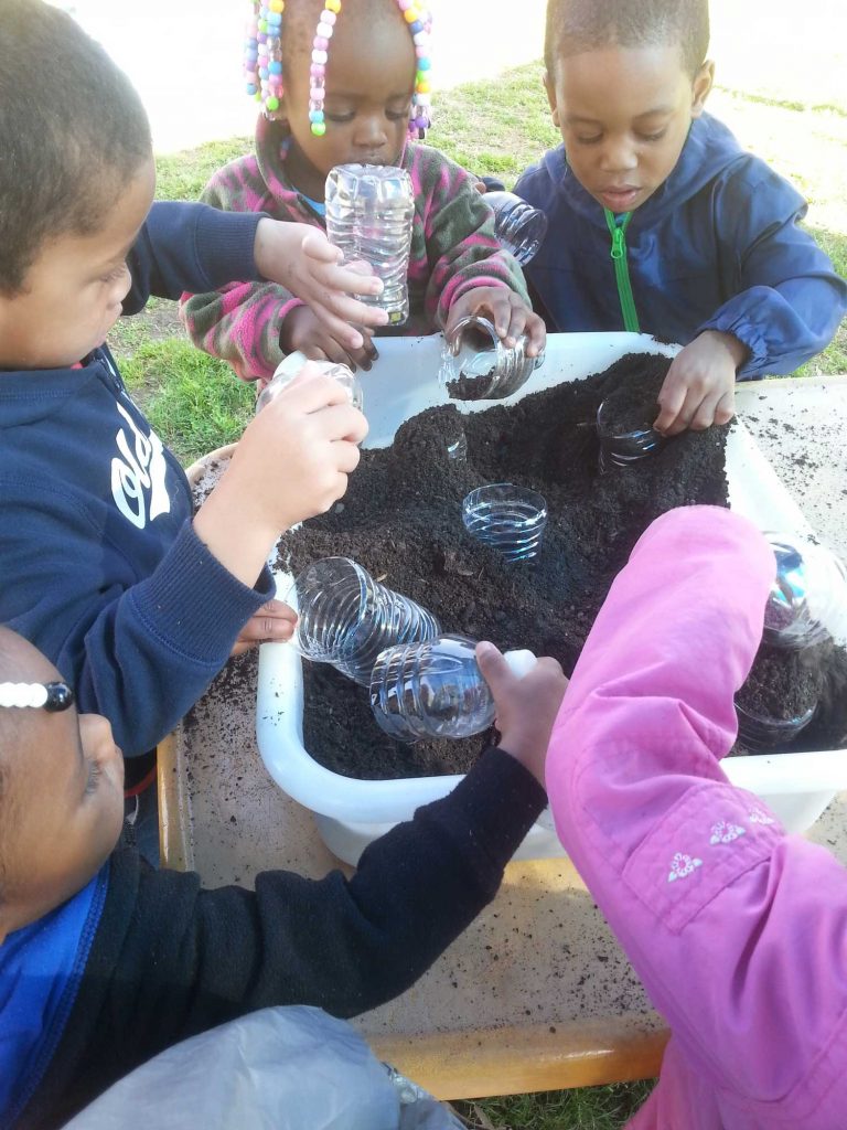 A group of children engaged in outdoor planting activities, with black soil and half-cut water bottles